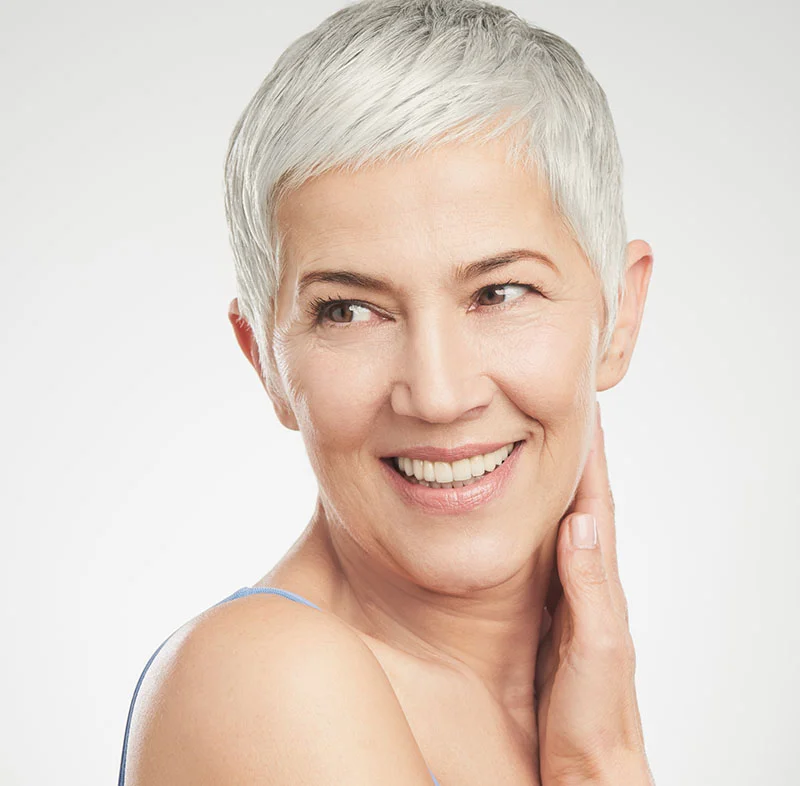 An older woman with short silver hair smiles while touching her neck. She is wearing a light blue tank top and has a light, neutral background - Dental Implant Restoration in Fort Lee, NJ