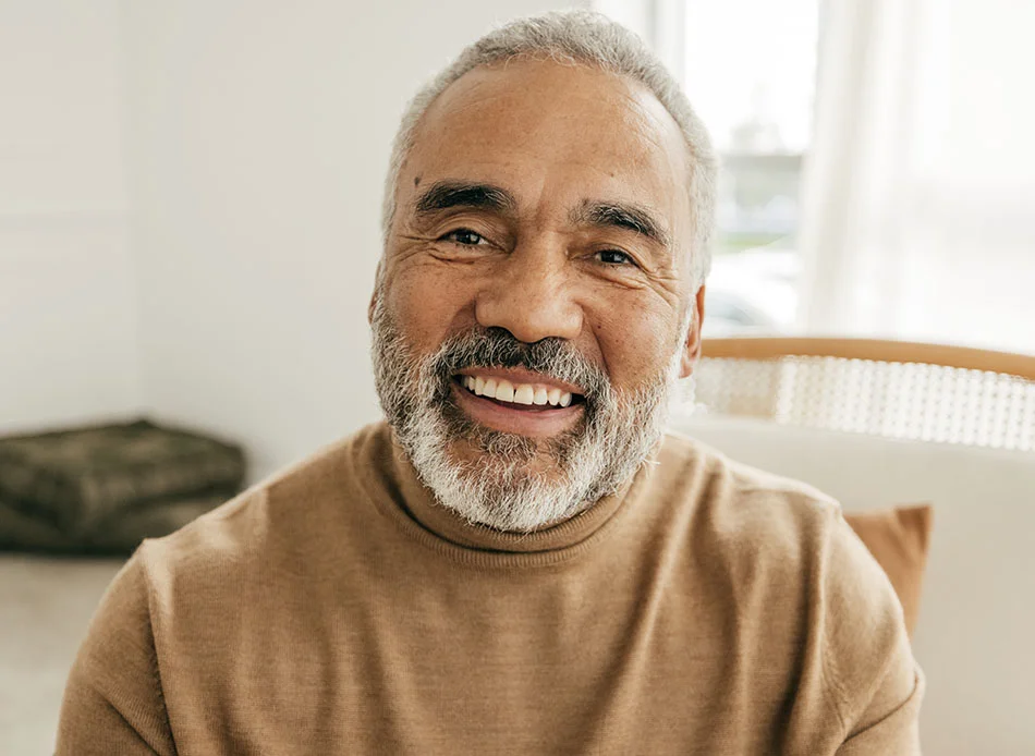 An older man with a gray beard, smiling and sitting indoors, wearing a brown sweater - Elder Care in Fort Lee, NJ