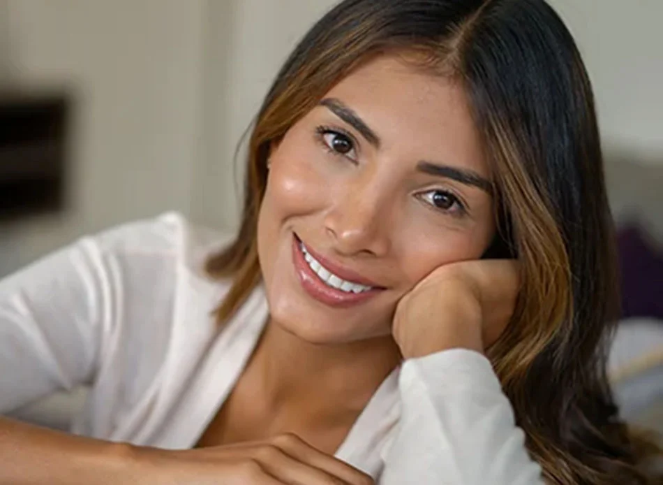 A relaxed young woman with long, dark hair, leaning her head on her hand, smiling warmly, and wearing a light-colored top - Injectables in Fort Lee, NJ