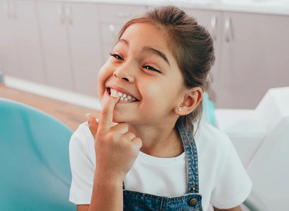 A young girl in overalls and a white shirt pointing at her tooth and smiling while sitting in a dental chair - Pediatric Dentistry in Fort Lee, NJ