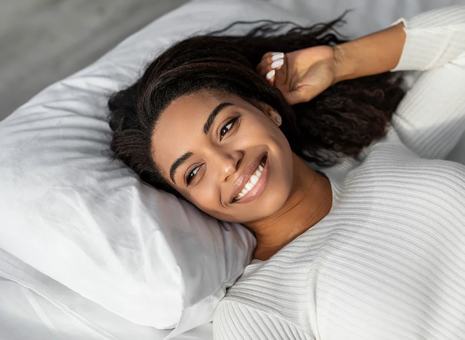 A woman with curly dark hair smiles happily while reclining on a pillow. She wears a light-colored ribbed top and is resting her head on her hand in a relaxed manner - Mandibular Advancement Devices in Fort Lee, NJ