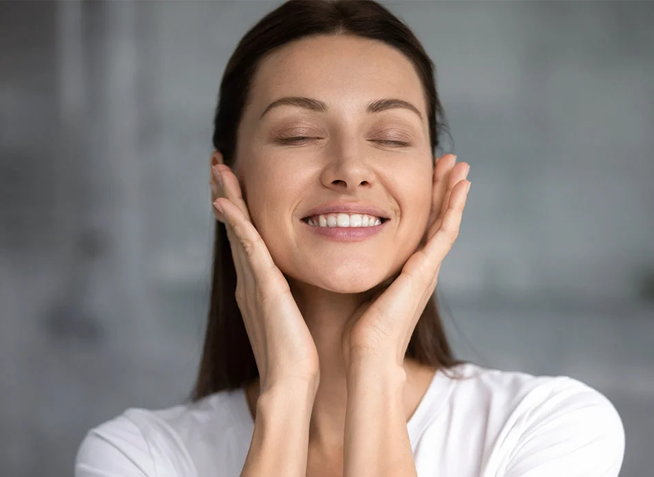 A woman with long brown hair smiles softly with her eyes closed while gently cradling her face with both hands. She appears relaxed and content, wearing a white top, in a calm and serene environment with soft, natural lighting - Scaling and Root Planing in Fort Lee, NJ